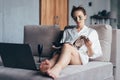 Young woman sitting at home with face beauty mask and reading magazine