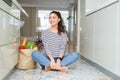 Young woman sitting on the kitchen floor with a paper bag full of fresh groceries looking away to side with smile on face, natural