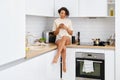 Young woman sitting on kitchen table top with mobile phone while waiting for the coffee to be brewed Royalty Free Stock Photo