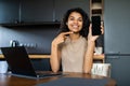 Young woman sitting at kithen with laptop pointed on phone screen