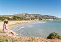 Young woman sitting and looking at a panoramic view of a sandy beach on the Mediterranean coast.