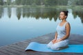 A young woman is sitting loosely on a mat by the lake holding a religious rosary with her eyes closed. Meditation in nature