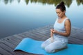 A young woman is sitting loosely on a mat by the lake holding a religious rosary. Yoga class in nature