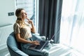 Young smiling woman is sitting on modern chair near the window in light cozy room at home working on laptop in relaxing atmosphere Royalty Free Stock Photo