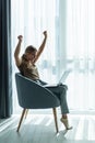 Young smiling woman is sitting on modern chair near the window in light cozy room at home working on laptop in relaxing atmosphere Royalty Free Stock Photo