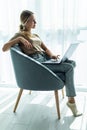 Young smiling woman is sitting on modern chair near the window in light cozy room at home working on laptop in relaxing atmosphere Royalty Free Stock Photo