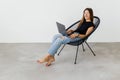 Young smiling woman is sitting on modern chair near the window in light cozy room at home working on laptop in relaxing atmosphere Royalty Free Stock Photo