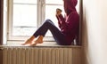 Young woman sitting near the window looking outside drinking coffee in a nostalgic mood