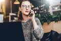 Young woman sitting in office, cafe at table in front of laptop and talking on mobile phone. Freelancer works in a cafe. Royalty Free Stock Photo