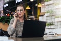 Young woman sitting in office, cafe at table in front of laptop and talking on mobile phone. Freelancer works in a cafe. Royalty Free Stock Photo