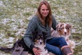 Young woman is sitting outside in the woods with her two Australian Shepherd dogs. Snow on the grass, twilight by the campfire.