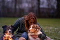 Young woman is sitting outside in the woods with her two Australian Shepherd dogs. Snow on the grass, twilight by the