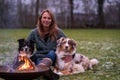 Young woman is sitting outside in the woods with her two Australian Shepherd dogs. Snow on the grass, twilight by the campfire.