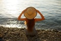 Young woman is sitting on the pebble shore with beach dress on holding a straw hat on her head. Back view portrait of