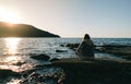 Young woman sitting on a rock, gazing in the distance, looking in the sea.