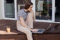 Young woman sitting slouching outdoor on terrace, working remotely online on laptop and drink fresh juice side view