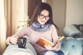 Young woman is sitting on a sofa and reading a book while holding a cup of coffee or tea Royalty Free Stock Photo