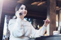 Young woman sitting at table in cafe, talking on cell phone, taking notes in notebook, working on laptop. Girl calls her friends Royalty Free Stock Photo