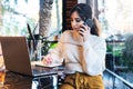 Young woman sitting at table in cafe, talking on cell phone, taking notes in notebook, working on laptop. Girl calls her friends Royalty Free Stock Photo