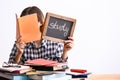 Young woman sitting at table with a lot of books and holding chalkboard with word STUDY against white background