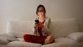 Young woman is sitting on white sofa looking into the smartphone with yellow amber blue light blocking eye glasses Royalty Free Stock Photo