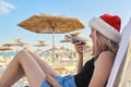 Young woman with smartphone in Santa hat of Claus resting on beach in sun lounger Royalty Free Stock Photo
