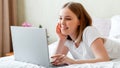 Young woman smile and work using laptop while lying in bed in morning at home. Happy girl in pajamas studying online or planning Royalty Free Stock Photo