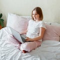Young woman smile and work using laptop while sitting in bed at morning home. Happy girl in pink pajamas studying online shopping Royalty Free Stock Photo