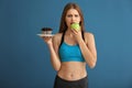 Young woman in sportswear choosing between cake and apple on color background Royalty Free Stock Photo