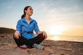 A young woman in sportswear is sitting cross-legged on the sand. In the background, the sea and sunset. Bottom view. Copy space. Royalty Free Stock Photo