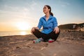 A young woman in sportswear is sitting cross-legged on the sand. In the background  the sea and sunset. Bottom view. Wellness and Royalty Free Stock Photo