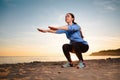 A young woman in sportswear trains by doing squats. In the background, sunset, sandy beach and ocean. Bottom view Royalty Free Stock Photo