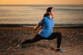 A young woman in sportswear trains by performing a leg stretch. In the background, sunset, sandy beach and ocean. Side Royalty Free Stock Photo