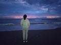 Young woman standing alone from behind on the beach, watching the sunset with dramatic blue sky clouds over the sea horizon.. Royalty Free Stock Photo