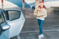 Young woman standing alone next to her car in the mall parking, with full hands of Christmas gifts