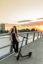 A young woman is standing by the bridge fence while talking on her phone with her electric scooter parked Royalty Free Stock Photo