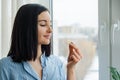 Young woman standing at home near the window taking vitamin Omega 3 fish oil yellow capsule