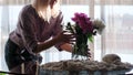 A young woman standing in a modern cozy living room puts a vase with a bouquet of beautiful fresh lilac peonies on the Royalty Free Stock Photo