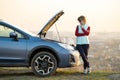 Young woman standing near broken down car with popped up hood having trouble with her vehicle. Female driver waiting for help Royalty Free Stock Photo