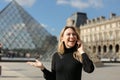 Young woman standing near Louvre and glass pyramind in black dress in Paris, talking by smartphone.