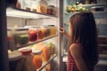 A young woman standing near an open fridge full of healthy food, vegetables, and fruits. A young girl full rear view looking at Royalty Free Stock Photo