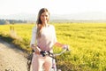 Young woman standing next to her bike over dusty country road, strong afternoon sun backlight in background shines on yellow