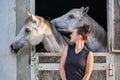 Young woman standing next to stable box looking to two white Arabian horses inside Royalty Free Stock Photo