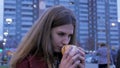 Young woman standing in an urban street and eating burger. Young woman eating fast food standing on the street