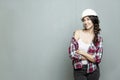 A young woman stands in front of a painted gray wall. A beautiful brunette in a construction helmet and a plaid shirt. Repair and Royalty Free Stock Photo