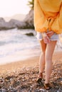 Young woman stands on a pebble beach with her hands folded behind her back. Back view. Cropped Royalty Free Stock Photo