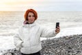 Young woman stands on the pebble seashore against the sunset and takes a selfie