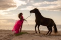 Young woman stands in a pink dress on a beach, with a horse beside her Royalty Free Stock Photo