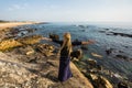 A young woman stands on the rocky shore of the ocean watching the surf