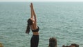 young woman stands in Urdhva Hastasana on a rock with the endless ocean below